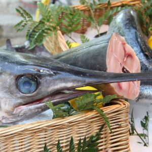 Plano directo a la cabeza de un pez espada fresco en una cesta junto a algunos limones. A su lado lo que parece ser la otra mitad del pez espada